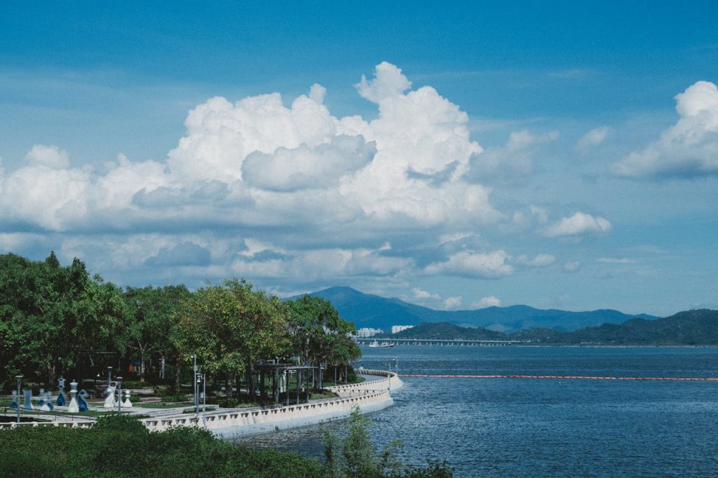A scenic coastal landscape featuring lush greenery, a clear blue sky, and distant mountains over a tranquil body of water.